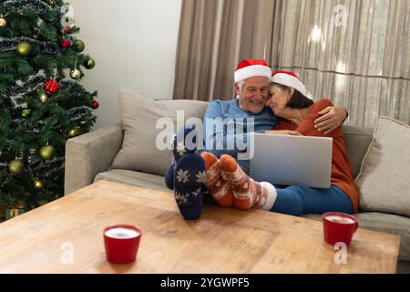 Couple senior dans les chapeaux de Père Noël câlin sur le canapé avec ordinateur portable par arbre de Noël, à la maison Banque D'Images