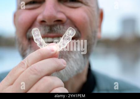Gros plan d'un homme mûr souriant largement, se concentrant sur sa main tenant une prothèse dentaire claire. Banque D'Images