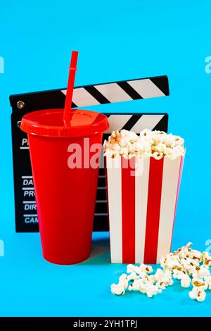 Image verticale de claquette de film derrière seau en carton rouge avec des rayures blanches avec pop-corn et tasse de soda rouge sur fond bleu, concept de nourriture Banque D'Images