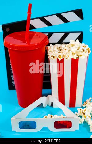 Image verticale de claquette de film derrière un verre de soude rouge, seau de pop-corn et lunettes 3D sur un fond bleu, concept de divertissement de cinéma Banque D'Images