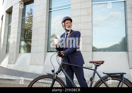 Responsable senior déterminé tenant une tasse de café et regardant loin tout en marchant à vélo dans la rue Banque D'Images