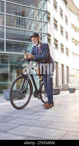 Homme d'affaires senior dans la messagerie texte de casque sur téléphone intelligent tout en se tenant debout avec le vélo sur la rue de la ville Banque D'Images