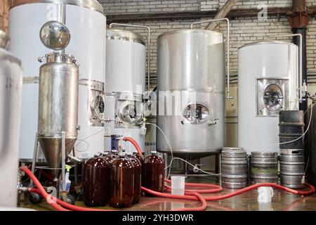 Plusieurs réservoirs en acier inoxydable stockant du cidre de pomme ou de la bière artisanale pendant le processus de fermentation en atelier, avec des fûts en plastique sur le sol prêts à être remplis Banque D'Images