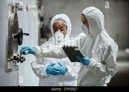 Vue latérale de la technicienne de processus féminine en combinaison Hazmat à l'aide d'un ordinateur tablette inspectant les réservoirs en acier inoxydable avec un collègue tout en contrôlant la fermentation p Banque D'Images