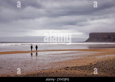 Deux femmes promènent un chien sous la pluie à travers le sable humide à marée basse à Saltburn Beach, Teesside, Angleterre. Banque D'Images