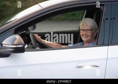 Une femme âgée joyeuse avec des lunettes est assise derrière le volant d'une voiture blanche, tenant le volant avec confiance pendant qu'elle aime sa conduite. Banque D'Images