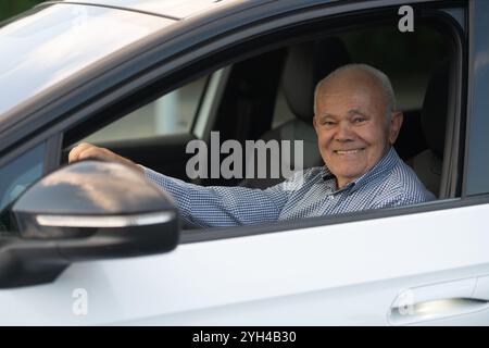 Un vieil homme joyeux dans une chemise à carreaux est assis dans le siège du conducteur d'une voiture blanche, souriant avec confiance et profitant du moment. Banque D'Images