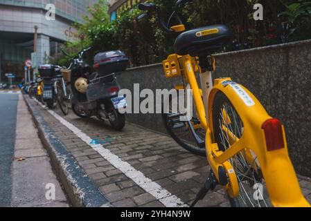 Une rangée de vélos et de scooters longe un trottoir à Shanghai, avec un vélo jaune issu d'un programme de partage. Un bâtiment moderne se dresse dans le backgroun Banque D'Images