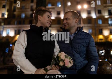 Un couple se promène dans une ville la nuit, la femme tenant un bouquet de fleurs Banque D'Images