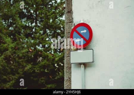 Panneau de signalisation routière monté sur un mur avec un cercle rouge et un fond bleu indiquant une interdiction des virages à gauche et à droite. Ci-dessous, un panneau blanc vide offre Banque D'Images