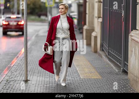 Femme élégante en Bourgogne Coat Walks City Street. Porter des vêtements à la mode chandail blanc à col roulé, collier de perles et Jeans gris, portant blanc Banque D'Images