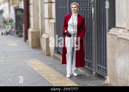 Femme mature élégante dans la ville à la mode Bourgogne manteau Standing City, portant des vêtements à la mode pull à col roulé blanc, collier de perles et Jeans gris. CONF Banque D'Images