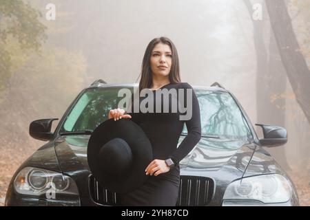 Femme élégante en robe noire et chapeau posant par un SUV noir dans un paysage d'automne Foggy. Banque D'Images