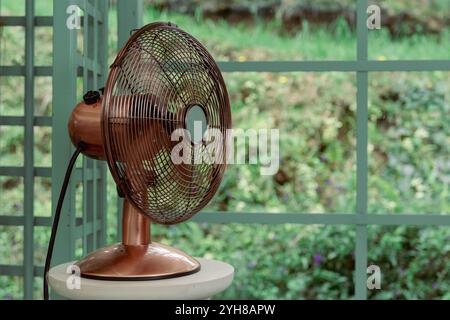 Un ventilateur électrique de couleur cuivre se trouve sur un piédestal blanc devant un fond vert et flou. Le ventilateur a une cage métallique ronde et est connecté à un Banque D'Images