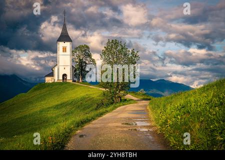 Route rurale sale courbe et église alpine emblématique sur la crête de la montagne, église Saint Primoz, village de Jamnik, Slovénie, Europe Banque D'Images