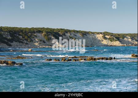 Les eaux tranquilles reflètent la lumière du soleil tandis que les vagues douces rencontrent des formations rocheuses près du rivage à midi. Banque D'Images