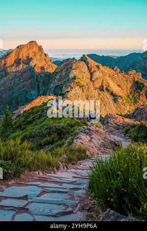 Une vue panoramique sur un sentier de montagne avec des randonneurs, entouré d'une végétation luxuriante, illuminé par la lumière dorée du soleil. Banque D'Images