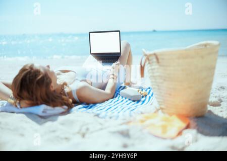 femme élégante détendue sur le bord de mer avec sac de paille, écran blanc d'ordinateur portable et serviette rayée. Banque D'Images