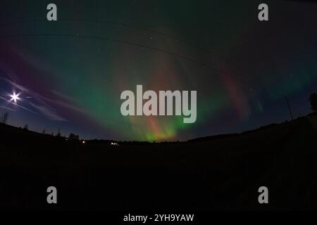 Les aurores boréales illuminent le ciel au-dessus d'une ferme rurale, avec la pleine lune brillante et des lignes électriques coupant le paysage serein. Banque D'Images