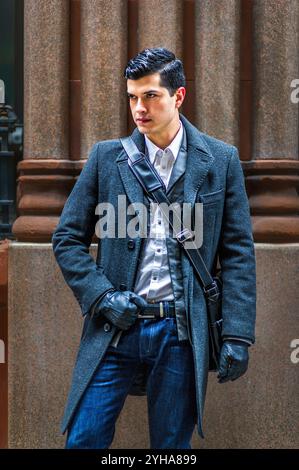 Un homme bien habillé se tient confiant devant une colonne de pierre, portant un manteau sombre et des gants avec un sac à bandoulière. Sa position et son style serrés Banque D'Images