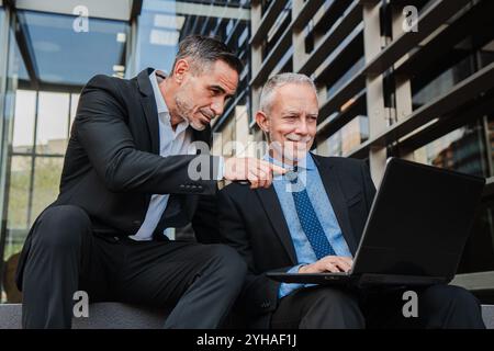 Deux hommes d'affaires expérimentés seniors en costumes formels discutant de la stratégie du projet, analysant les données commerciales ensemble sur ordinateur portable à l'extérieur. Cadres matures utilisant un ordinateur sur le lieu de travail de bâtiment d'entreprise. Photo de haute qualité Banque D'Images