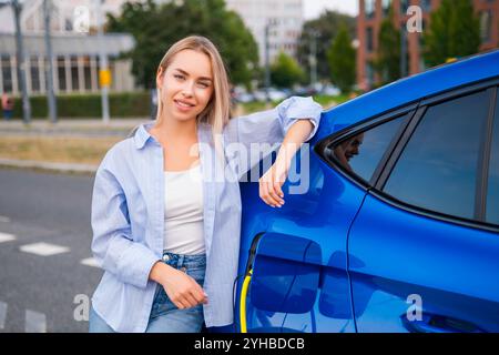 Femme blonde sourit tout en se relaxant à côté de son véhicule électrique alors qu'il se recharge dans la rue Banque D'Images