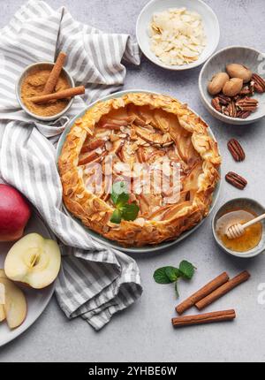 Une tarte aux pommes magnifiquement présentée ornée d'amandes et de miel se trouve sur une table rustique. Autour de lui se trouvent des pommes fraîches, des bâtonnets de cannelle et des bols Banque D'Images