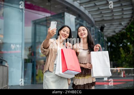 Deux jolies amies asiatiques heureuses aiment prendre des selfies avec un smartphone après leur virée shopping au centre commercial, montrant leurs achats Banque D'Images