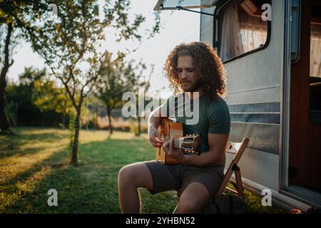 Jeune homme est en voyage de camping dans la nature seul, assis dans la caravane devant et jouant de la guitare. Banque D'Images