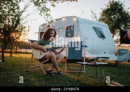 Jeune homme est en voyage de camping dans la nature seul, assis dans la caravane devant et jouant de la guitare. Banque D'Images