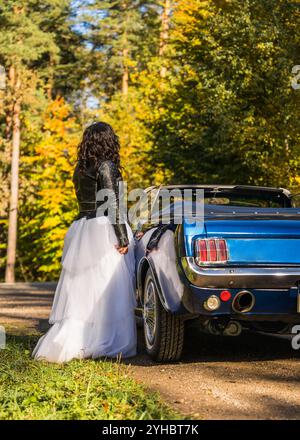 Femme à la mode portant une veste en cuir noir, longue jupe bouffante blanche, bottes en cuir se tient à côté de la voiture bleue dans une forêt d'automne. Son visage est caché b Banque D'Images