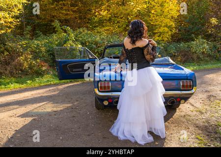 Femme à la mode portant une veste en cuir noir, longue jupe bouffante blanche, bottes en cuir se tient à côté de la voiture bleue dans une forêt d'automne. Son visage est caché b Banque D'Images