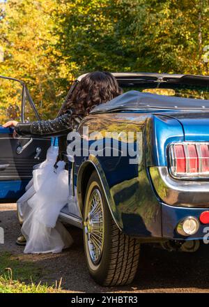 Femme à la mode portant une veste en cuir noir, longue jupe bouffante blanche et bottes en cuir se trouve dans une voiture bleue vintage dans une forêt d'automne. Vue arrière Banque D'Images