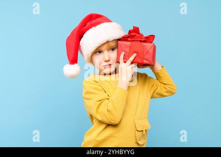 Garçon mignon portant le chapeau rouge santa tenant le cadeau de noël enveloppé, portrait de studio isolé sur fond bleu pastel. Arrière-plan des fêtes. Banque D'Images