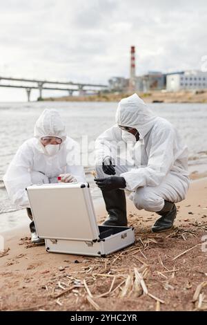 Plan vertical pleine longueur de deux scientifiques prenant des sondes dans la nature et portant des combinaisons de protection sur site industriel, espace copie Banque D'Images