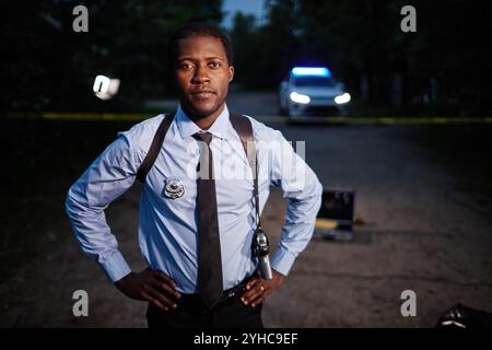 Portrait de taille haute d'un jeune homme afro-américain en tant qu'officier de police portant l'uniforme debout sur la scène de crime la nuit et regardant la caméra, l'espace de copie Banque D'Images