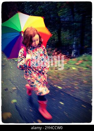 A young redheaded girl hurries to school on a rainy day wearing a floral rain coat red boots and carrying a rainbow umbrella - Stock Photo