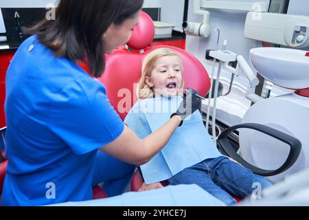 Gros plan d'un garçon mignon d'âge élémentaire assis dans le fauteuil du dentiste, ouvrant la bouche large pour le médecin méconnaissable avec la main dans le gant dans le cadre seulement en utilisant le den Banque D'Images