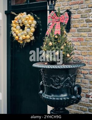 Une couronne décorative faite de boules d'or et d'argent avec des paillettes et des rubans suspendus sur une porte vert foncé aux pays-Bas avec un petit sapin de Noël Banque D'Images