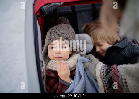 La jeune famille profite des vacances d'hiver dans les montagnes. Parents poussant les enfants dans la poussette de wagon à travers la forêt enneigée. Banque D'Images