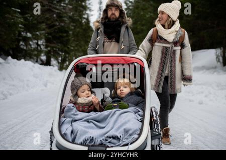 La jeune famille profite des vacances d'hiver dans les montagnes. Parents poussant les enfants dans la poussette de wagon à travers la forêt enneigée. Banque D'Images