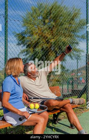 vertical deux joueurs de tennis seniors sont assis sur un banc près du court, partageant un moment joyeux alors qu’ils prennent un selfie avec un smartphone lors d’une pause Banque D'Images