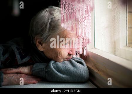 Une femme âgée regarde tristement par la fenêtre de sa maison, ses yeux reflétant un profond sentiment de mélancolie. Banque D'Images