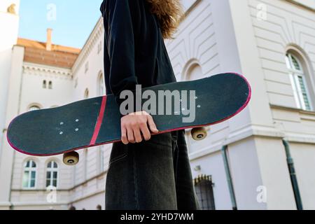 Skateboarder dans un Jean baggy tenant un skateboard rayé marchant dans la rue de la ville. Adolescent fait du skateboard. Concept de sous-culture de la jeunesse Banque D'Images