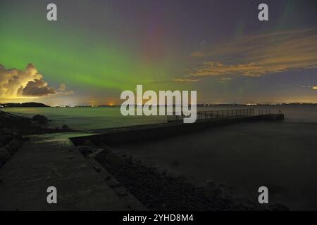 Aurores boréales au-dessus d'une mer avec lumières de ville lointaines et ciel partiellement nuageux, plage, Toensberg, Oslofjord, Norvège, Europe Banque D'Images