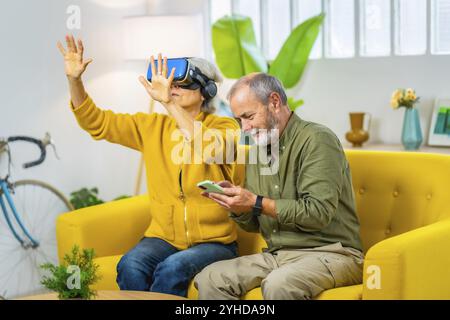 Femme âgée utilisant des googles de réalité virtuelle à côté de son mari assis sur le canapé à l'aide d'un téléphone portable Banque D'Images