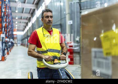 Ouvrier manuel masculin mature portant un gilet réfléchissant de sécurité à l'aide d'un chariot élévateur pour déplacer des boîtes dans un entrepôt Banque D'Images