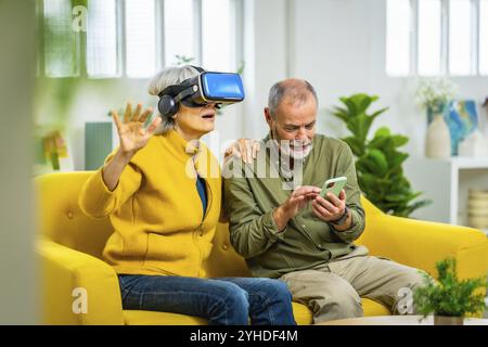 Femme âgée utilisant des lunettes de réalité virtuelle à côté de son mari assis sur le canapé à l'aide de l'application mobile Banque D'Images