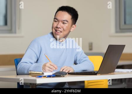 Un jeune étudiant universitaire asiatique faisant des travaux scolaires dans un bureau de classe heureux, écrivant des notes avec un crayon et son ordinateur portable, regardant vers d'autres peop Banque D'Images