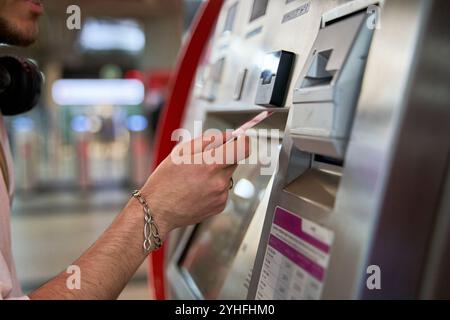 Une vue rapprochée capture un homme à la gare, insérant une carte ou de l'argent liquide pour acheter un billet, préparant son voyage et embrassant l'excitation Banque D'Images